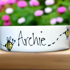 White pet bowl with 'Archie' and bee designs on a wooden surface with a blurred floral background
