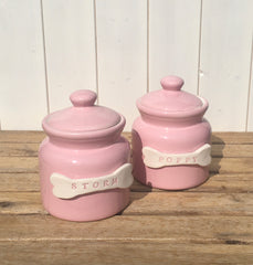 Two pink ceramic jars with lids labeled 'Storm' and 'Poppy' on a wooden surface.