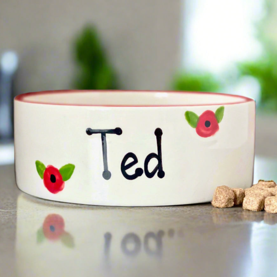 Ceramic pet bowl with 'Ted' and flower designs on a reflective surface.